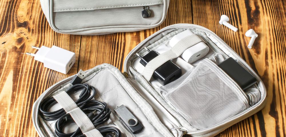 Open travel organizer with cables, chargers, and accessories on a wooden surface. Nearby are two wireless earbuds. Neat and organized presentation.