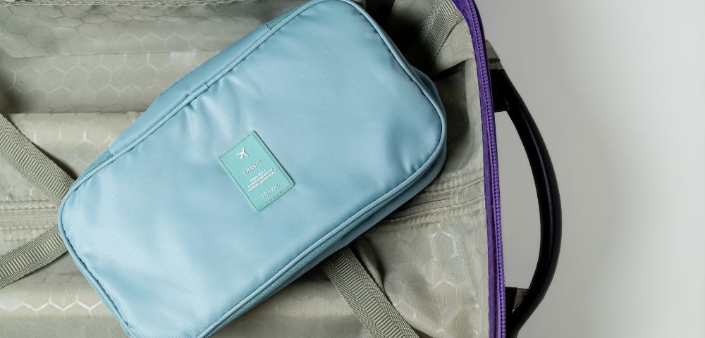 Blue toiletry bag sits inside an open suitcase with a grey interior and purple rim. The setting conveys a sense of travel readiness.
