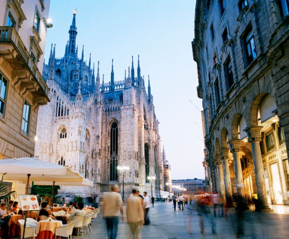 Street scene at dusk with Milan's Gothic cathedral towering on the left. People stroll by, and diners enjoy an outdoor cafe under umbrellas.