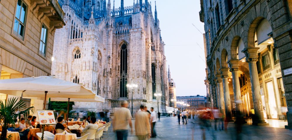 Street scene at dusk with Milan's Gothic cathedral towering on the left. People stroll by, and diners enjoy an outdoor cafe under umbrellas.