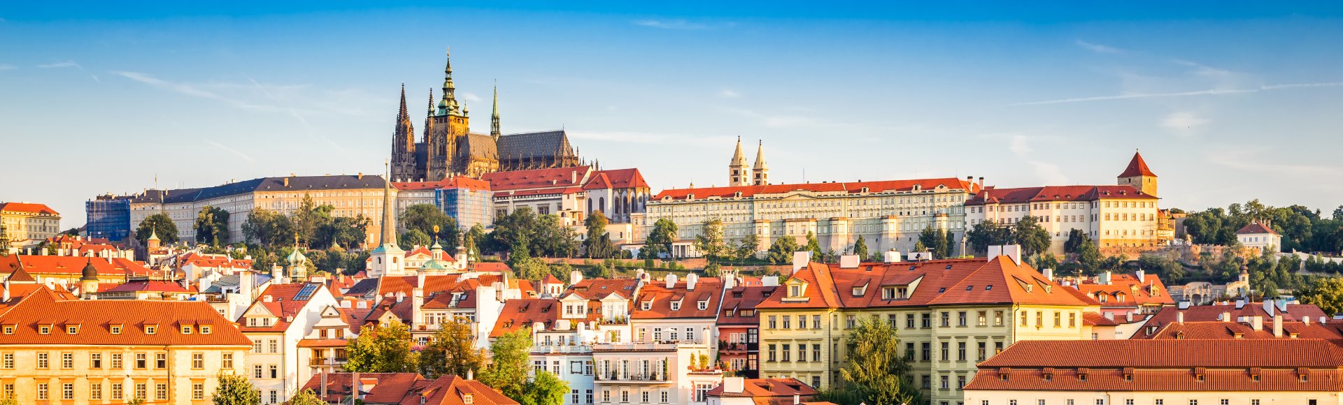 Panoramic view of Prague skyline featuring historic buildings with red roofs, dominated by a large castle under a clear blue sky, evoking a sense of history and grandeur.