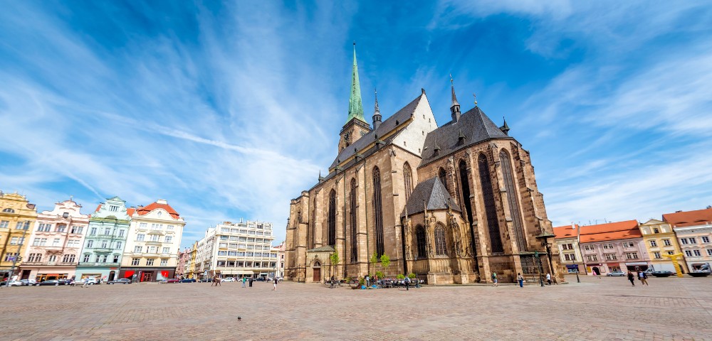 Historic cathedral with a tall spire in a vibrant town square, framed by colorful, ornate buildings. A few people wander under a bright blue sky.