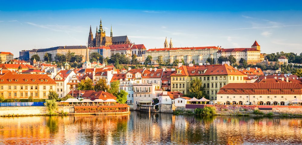 Panoramic view of Prague skyline featuring historic buildings with red roofs, dominated by a large castle under a clear blue sky, evoking a sense of history and grandeur.