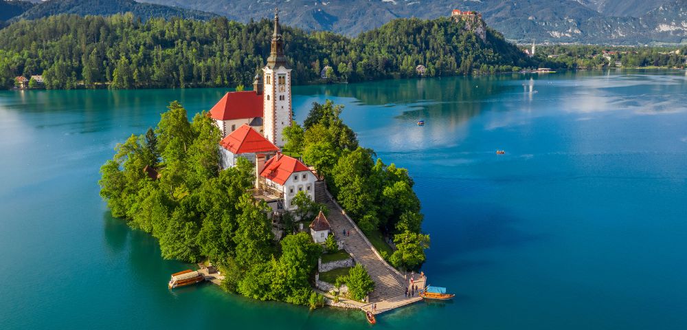 Scenic view of a small island with a church having a tall spire and red roofs, surrounded by a calm blue lake and lush green forests under a clear sky.