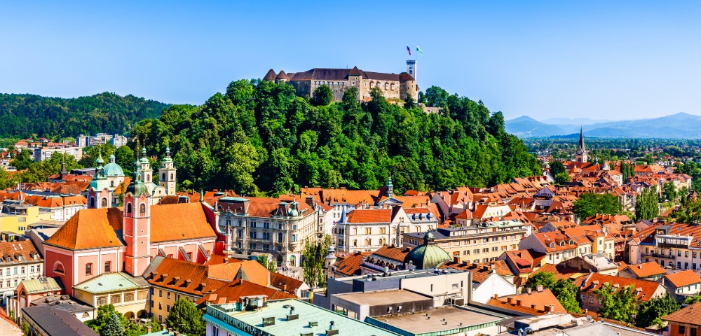Scenic view of Ljubljana, Slovenia, with a medieval castle atop a lush hill. Charming cityscape features red rooftops and historic architecture under a clear blue sky.