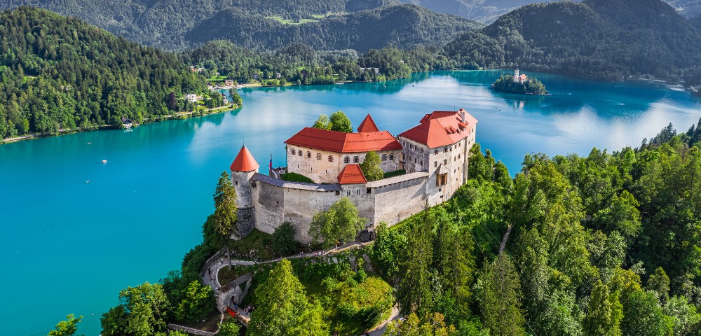 Aerial view of a historic castle with red roofs atop a forested hill, overlooking a serene turquoise lake surrounded by lush green mountains.