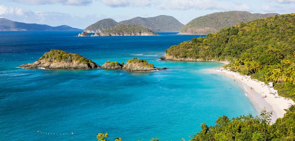 Turquoise waters surround small islands and a lush, green coastline in a tropical bay. A sandy beach curves along the shore, creating a serene and inviting scene.