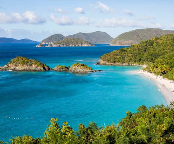 Turquoise waters surround small islands and a lush, green coastline in a tropical bay. A sandy beach curves along the shore, creating a serene and inviting scene.