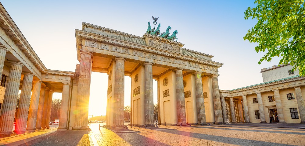 Brandenburg Gate at sunrise, bathed in golden light. Sunlight streams between the columns, casting long shadows. The tone is serene and majestic.