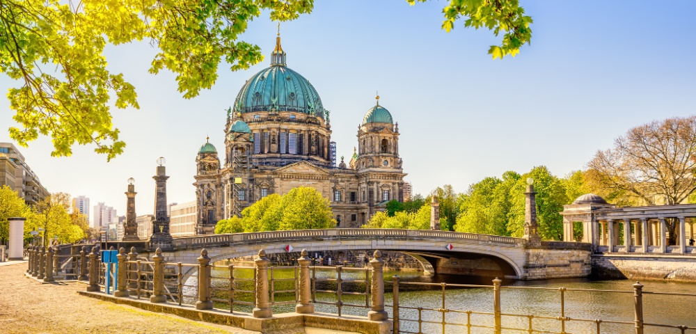 Berlin Cathedral with its large green dome stands majestically under a clear blue sky. A river and bridge are in the foreground, surrounded by lush trees.