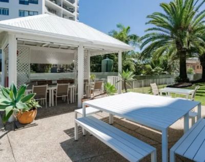 Outdoor dining area with a white gazebo, long tables, and benches. Palm trees and lush greenery surround the serene setting under a clear blue sky.