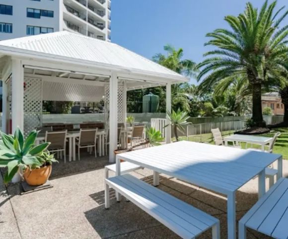 Outdoor dining area with a white gazebo, long tables, and benches. Palm trees and lush greenery surround the serene setting under a clear blue sky.