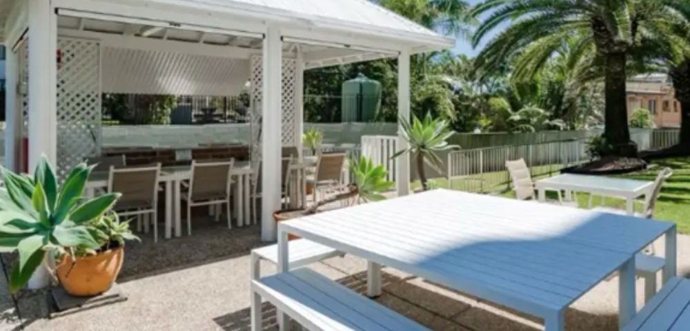 Outdoor dining area with a white gazebo, long tables, and benches. Palm trees and lush greenery surround the serene setting under a clear blue sky.