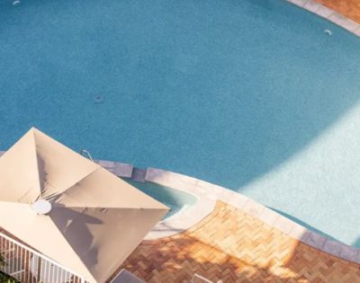 Aerial view of a serene, sunlit, oval-shaped swimming pool with clear blue water, bordered by neat pavers. Nearby is a beige patio umbrella.