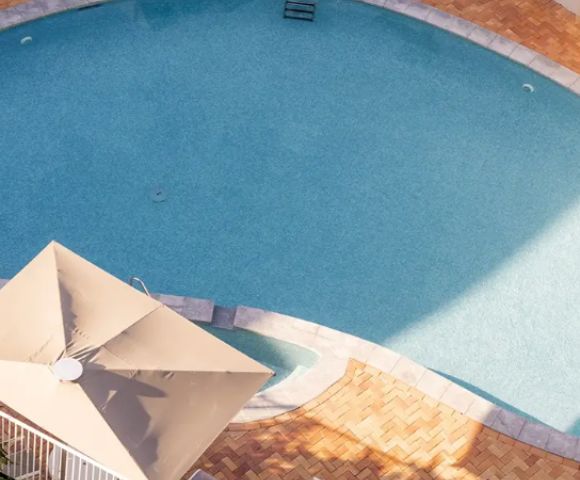 Aerial view of a serene, sunlit, oval-shaped swimming pool with clear blue water, bordered by neat pavers. Nearby is a beige patio umbrella.