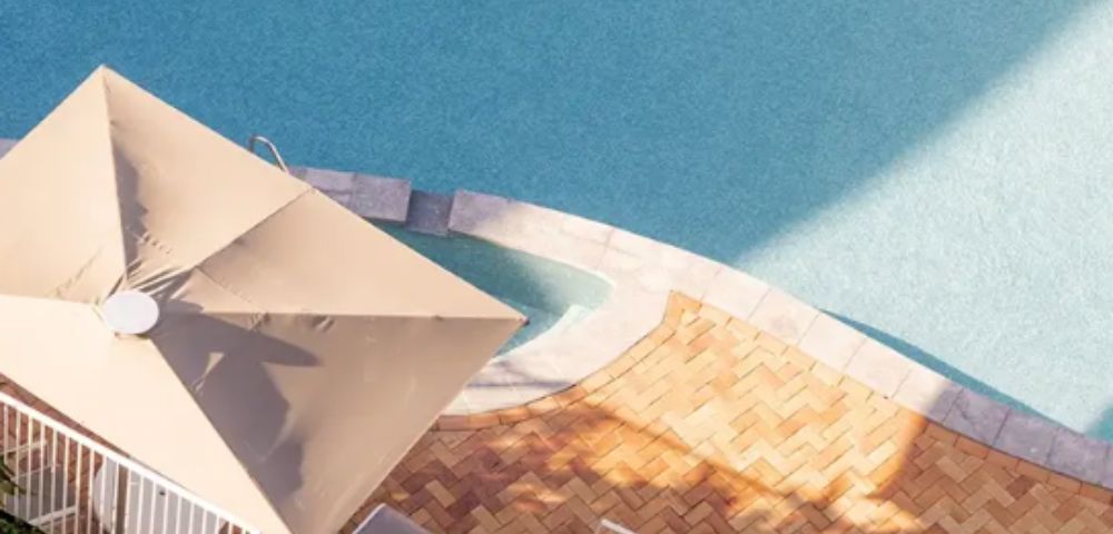 Aerial view of a serene, sunlit, oval-shaped swimming pool with clear blue water, bordered by neat pavers. Nearby is a beige patio umbrella.