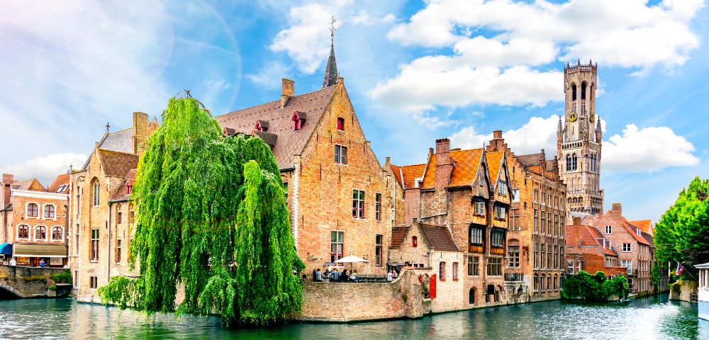 Historic canal-side buildings in Bruges with a large willow tree in the foreground and the Belfry tower in the background under a blue sky.