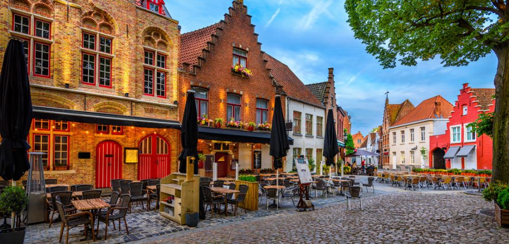 Quaint European square with cobblestone streets, colorful historic buildings, outdoor café seating, and a tree. The scene is peaceful and inviting.