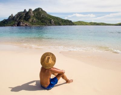A person in blue shorts and a straw hat sits on a sandy beach, gazing at a lush green island across the calm, blue ocean under a clear sky.