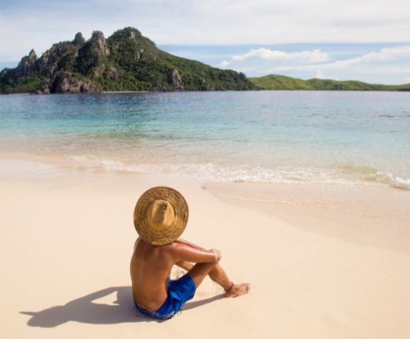 A person in blue shorts and a straw hat sits on a sandy beach, gazing at a lush green island across the calm, blue ocean under a clear sky.