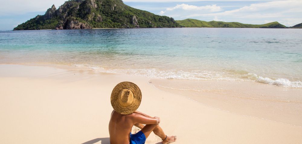 A person in blue shorts and a straw hat sits on a sandy beach, gazing at a lush green island across the calm, blue ocean under a clear sky.