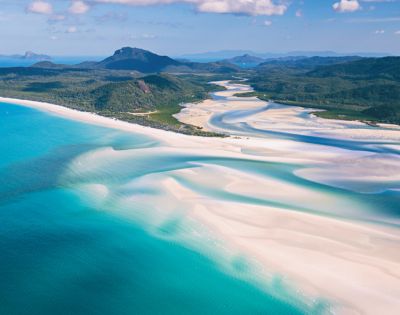 Aerial view of Whitehaven Beach, showcasing turquoise waters and swirling white sand patterns. Lush green hills and a blue sky create a serene landscape.