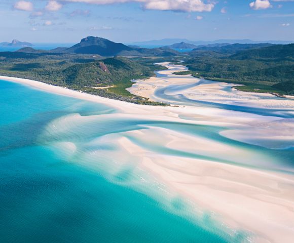 Aerial view of Whitehaven Beach, showcasing turquoise waters and swirling white sand patterns. Lush green hills and a blue sky create a serene landscape.