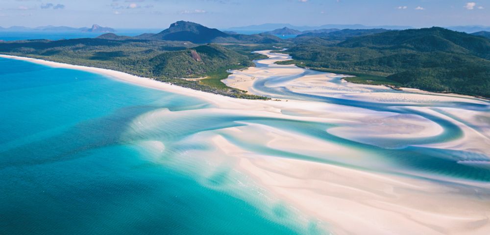 Aerial view of Whitehaven Beach, showcasing turquoise waters and swirling white sand patterns. Lush green hills and a blue sky create a serene landscape.