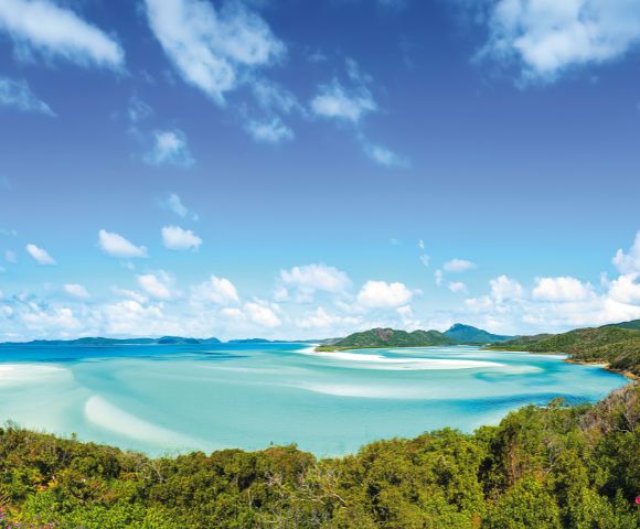 Expansive turquoise bay with swirling sands meets lush green forest under a bright blue sky dotted with fluffy clouds, evoking serenity and natural beauty.