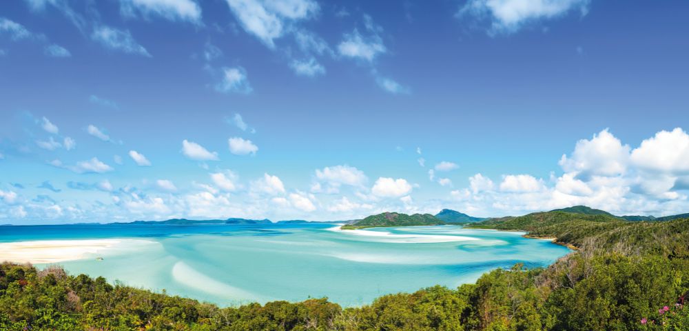 Expansive turquoise bay with swirling sands meets lush green forest under a bright blue sky dotted with fluffy clouds, evoking serenity and natural beauty.