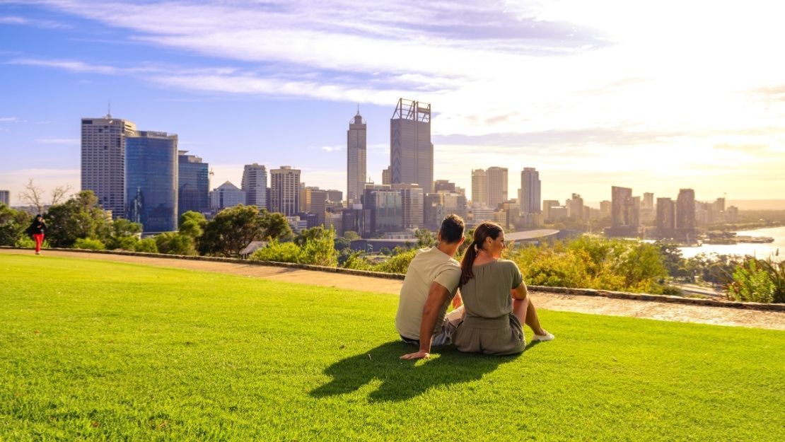 A couple sits on vibrant green grass, overlooking a city skyline under a blue sky. The scene conveys relaxation and a serene urban landscape.