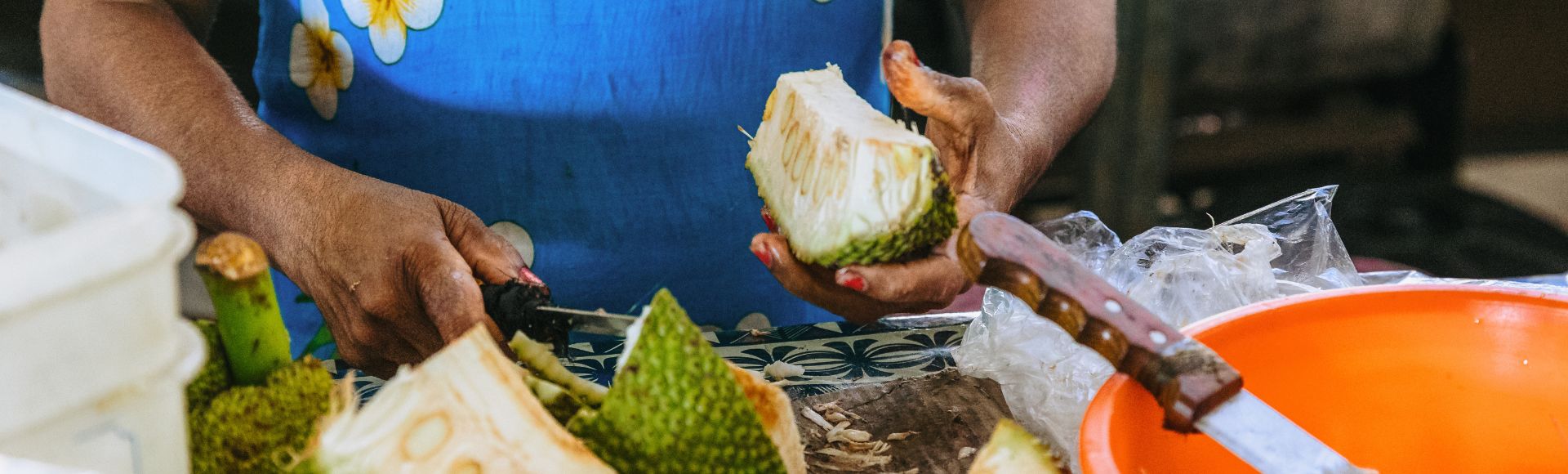 A person cuts a breadfruit with a knife. Slices are placed on a table with an orange bowl nearby, suggesting food preparation.
