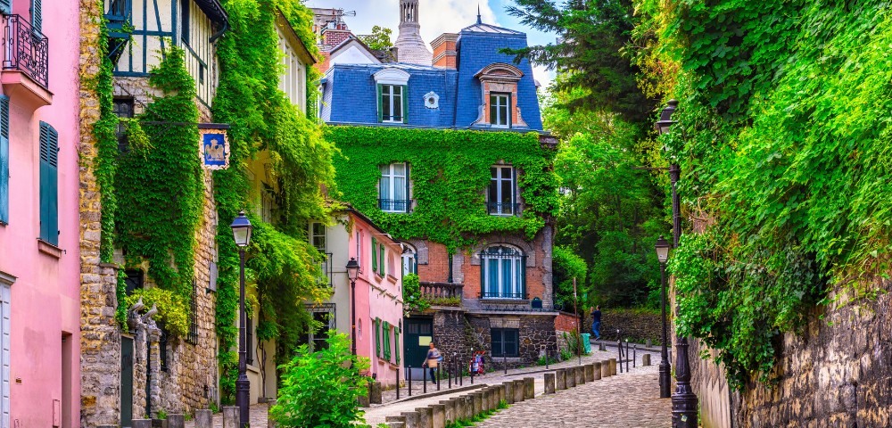 A quaint, cobblestone street lined with ivy-covered, colorful buildings in vibrant greens and pinks. The scene is cozy and picturesque, exuding charm.