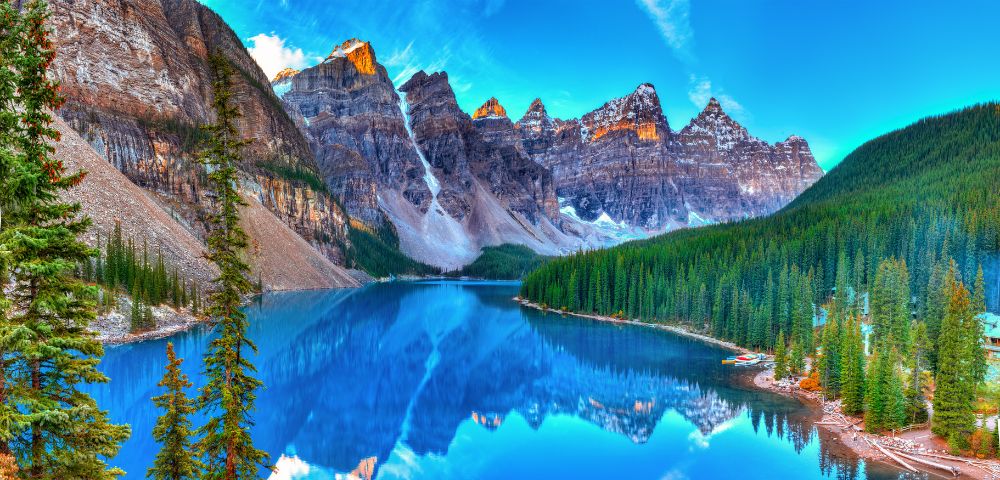 A serene mountain lake reflects rugged peaks and lush pines under a bright blue sky. The scene conveys tranquility and natural beauty.