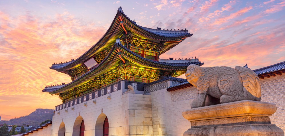 A traditional Korean palace gate under a vibrant sunset sky, adorned with intricate roof designs. A stone guardian statue stands in the foreground.