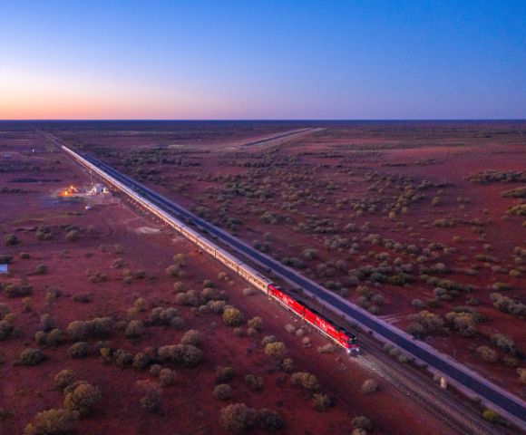 A red train travels through a vast, arid landscape during sunset. The sky is a gradient of warm colors, and scattered shrubs dot the reddish earth.