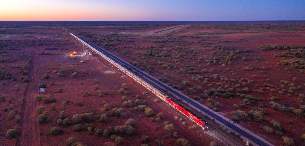A red train travels through a vast, arid landscape during sunset. The sky is a gradient of warm colors, and scattered shrubs dot the reddish earth.
