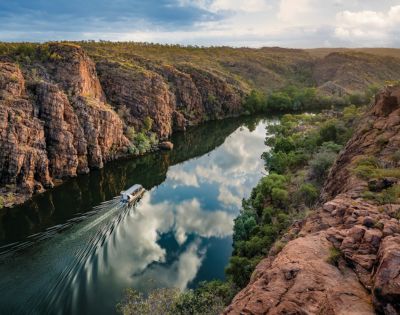 A boat travels along a narrow river reflecting clouds, surrounded by rugged cliffs and lush greenery under a partly cloudy blue sky, conveying serenity.