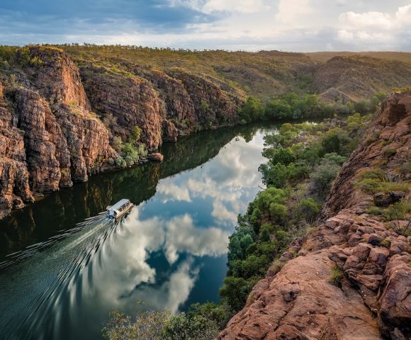 A boat travels along a narrow river reflecting clouds, surrounded by rugged cliffs and lush greenery under a partly cloudy blue sky, conveying serenity.