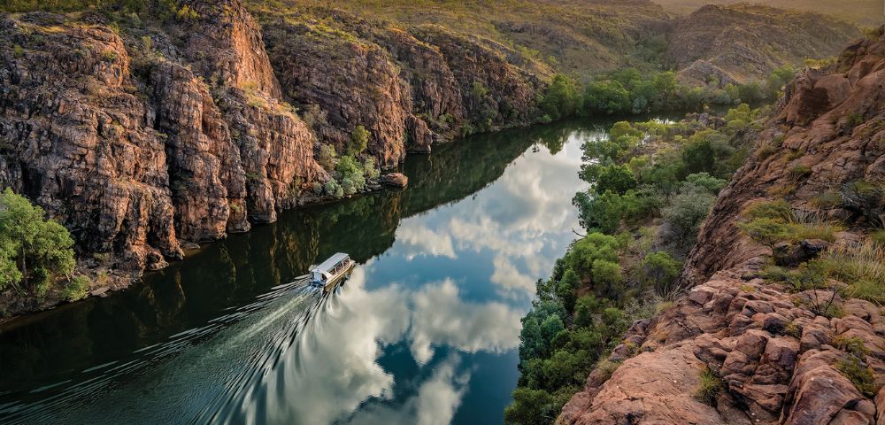 A boat travels along a narrow river reflecting clouds, surrounded by rugged cliffs and lush greenery under a partly cloudy blue sky, conveying serenity.