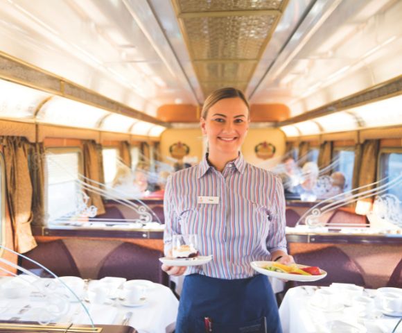 A smiling train stewardess in a striped shirt serves desserts on a luxury train with elegant table settings and plush seating, creating a warm, inviting atmosphere.