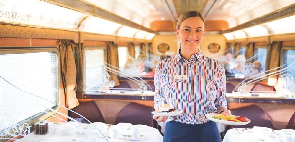 A smiling train stewardess in a striped shirt serves desserts on a luxury train with elegant table settings and plush seating, creating a warm, inviting atmosphere.