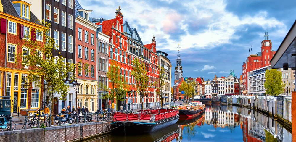 Colorful, traditional Dutch buildings line a canal in Amsterdam. Boats float on calm water reflecting the architecture under a partly cloudy sky.
