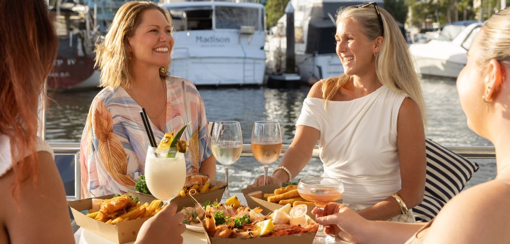 Two women smiling at an outdoor marina restaurant, sharing food and drinks. Sunlit boats in the background create a relaxed, cheerful vibe.