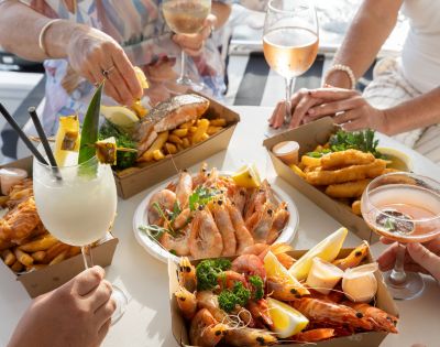 People enjoying a seafood feast with prawns, fish, chips, and cocktails at a sunlit table. The atmosphere is casual and cheerful, with hands reaching for food.
