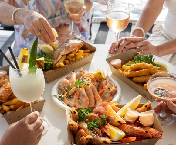People enjoying a seafood feast with prawns, fish, chips, and cocktails at a sunlit table. The atmosphere is casual and cheerful, with hands reaching for food.