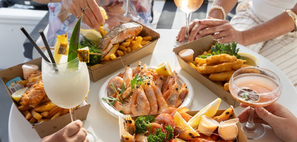 People enjoying a seafood feast with prawns, fish, chips, and cocktails at a sunlit table. The atmosphere is casual and cheerful, with hands reaching for food.