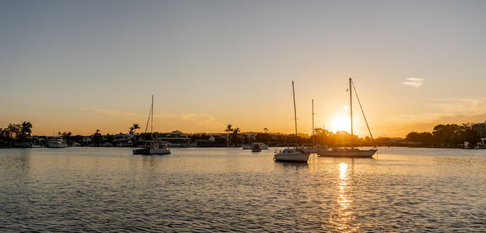Peaceful sunset over a harbor with silhouetted sailboats on calm water, casting reflections and creating a serene, golden-hued scene.