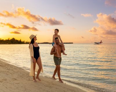 A family walks along a beach at sunset, with warm, golden sky hues. The child sits on the father's shoulders, and a small plane flies over the sea.