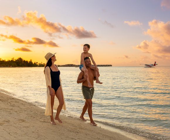 A family walks along a beach at sunset, with warm, golden sky hues. The child sits on the father's shoulders, and a small plane flies over the sea.